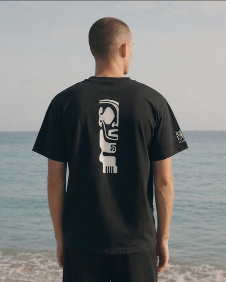 Homme de dos sur la plage portant le tee-shirt UVEA TIKI gris foncé, motif Tiki blanc au dos