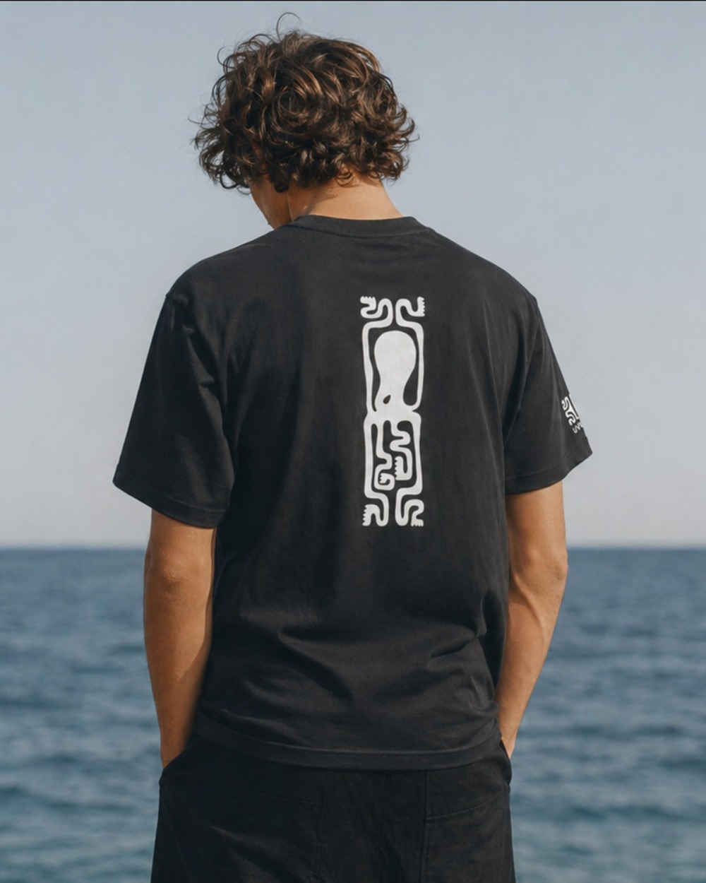 Hombre de espaldas en la playa con la camiseta UVEA OCTOPUS de algodón gris oscuro, pulpo tótem blanco en la espalda vue 1